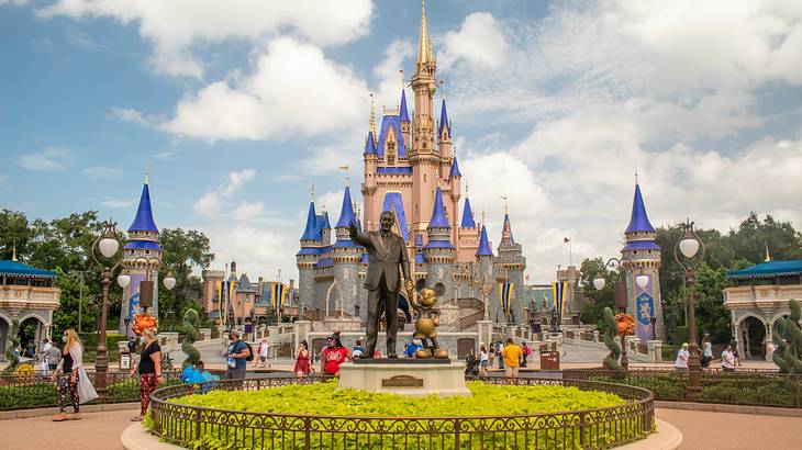 A statue of Walt Disney with Mickey Mouse against a castle under a partly cloudy sky