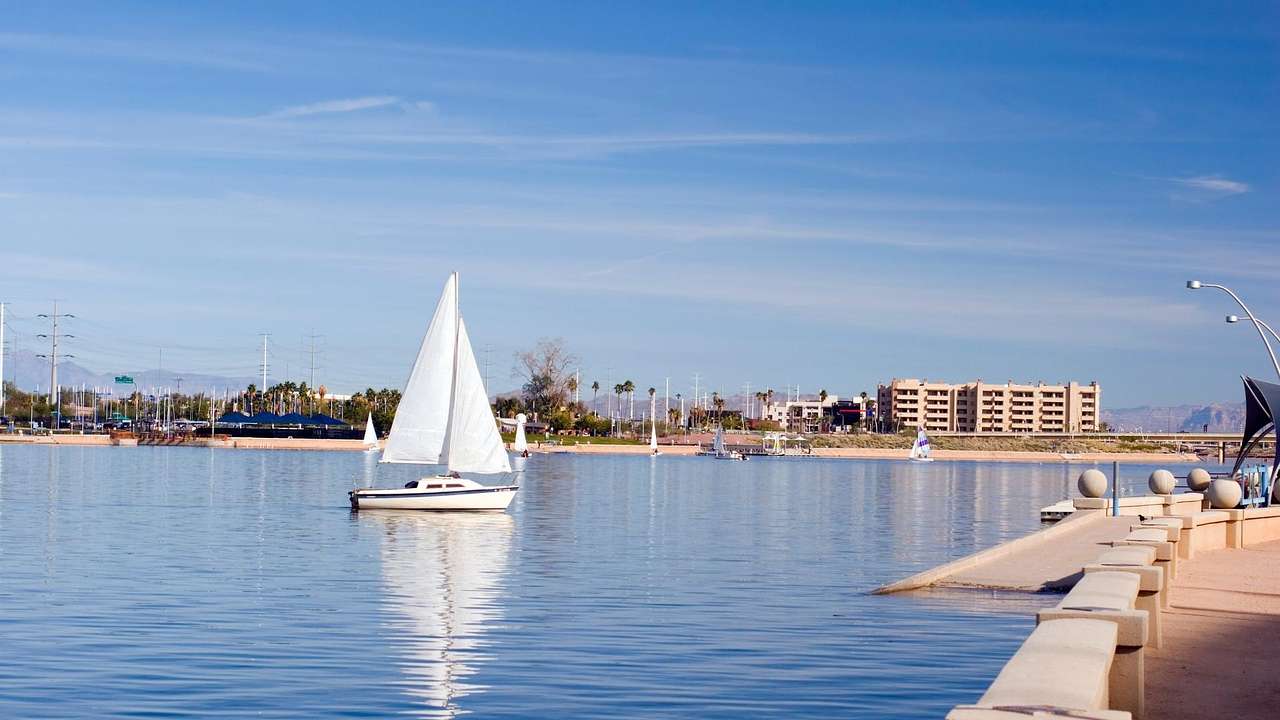 A sailboat on the water with buildings in the distance and a path to the side