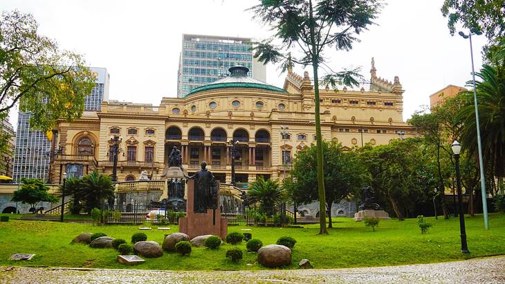 A stone building with arches and a domed roof next to a garden with a statue