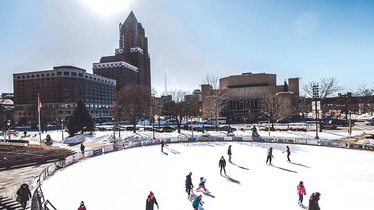 An ice rink with people skating on it and buildings behind it on a clear winter day