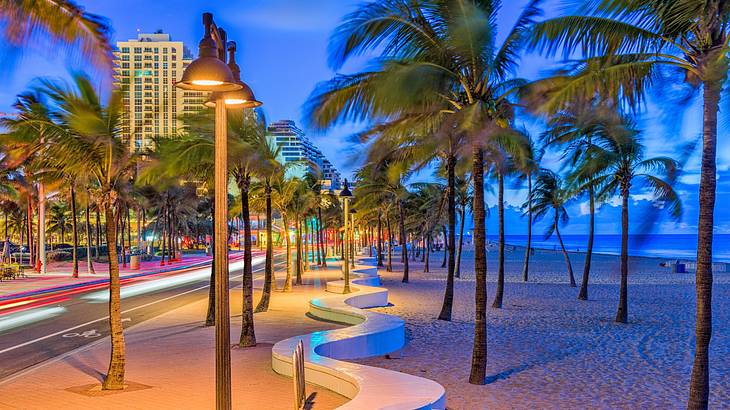 A street with lampposts next to a beach and palm trees at sunset