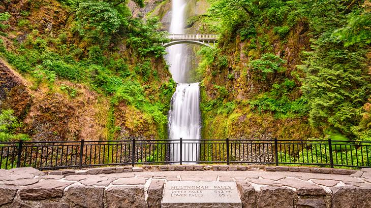 Stone signage by a black metal railing with a view of a stone bridge and a waterfall