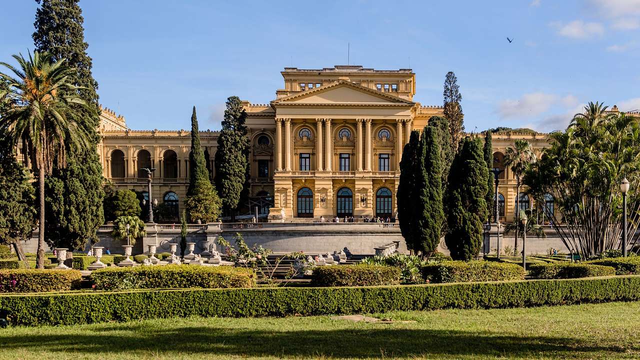 A large yellow-coloured stone building with columns next to a green garden and trees