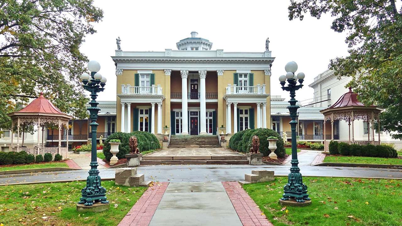 A mansion with columns, balconies, statues, and gazebos on the side next to a path