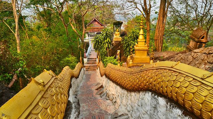 A long staircase with golden-scaled railings, a Buddha statue, and trees on the side