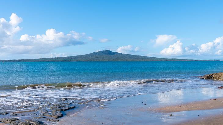 A mountain-like island next to the blue sea and a beach beneath a partly cloudy sky