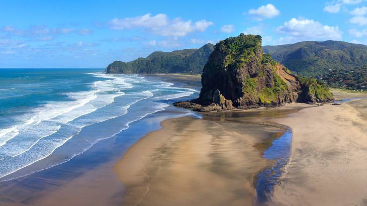 Blue sea with multiple rolling waves beside hills and the sand on a partly cloudy day