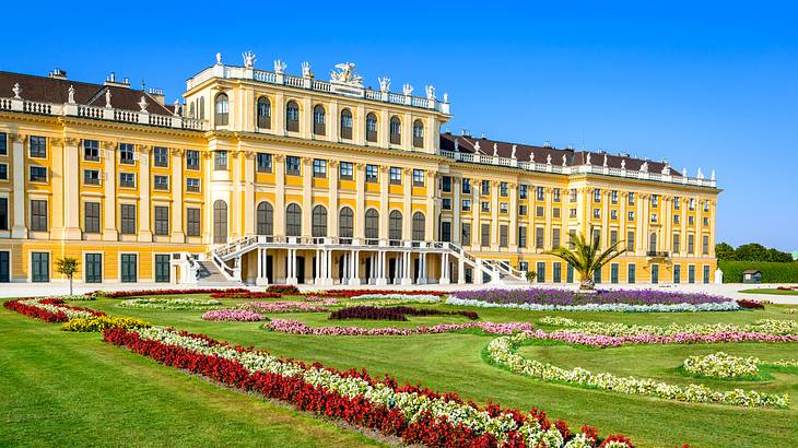 A large yellow-coloured palace with a lawn with red, pink, and white flowers