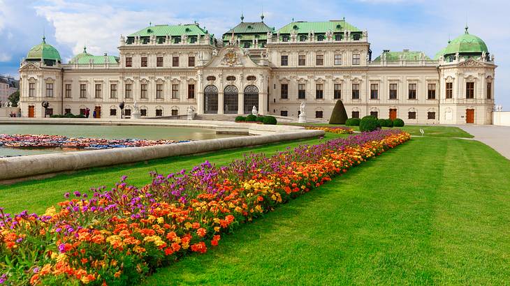 A large white palace next to a pond and colourful flowers on a manicured lawn