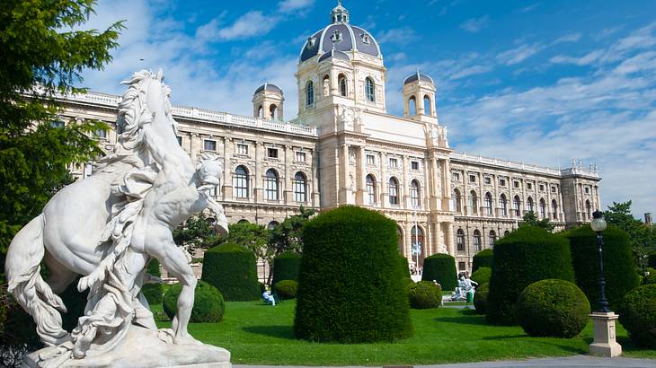 A large white building with a dome next to a lawn, hedges, and white statues