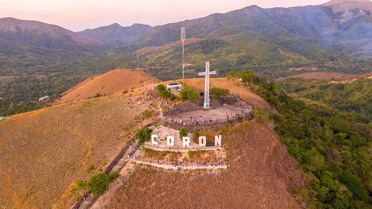 A hill with a cross sculpture and a sign that says "Coron," surrounded by other hills