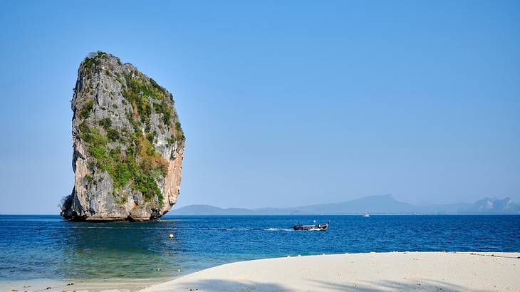 View of a big limestone rock in a body of water from a beach with a boat sailing by