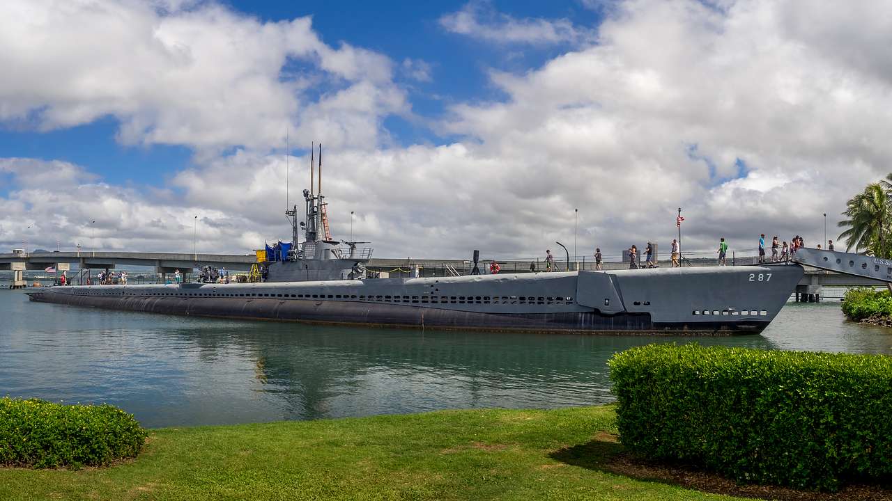 A gray submarine sitting above the water with grass and trees in front of it