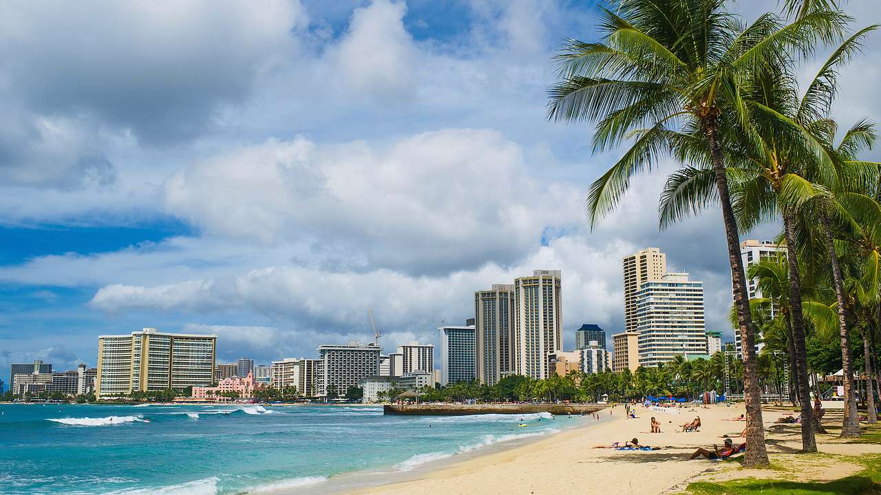 A sandy beach with palm trees, blue ocean, and buildings in the distance