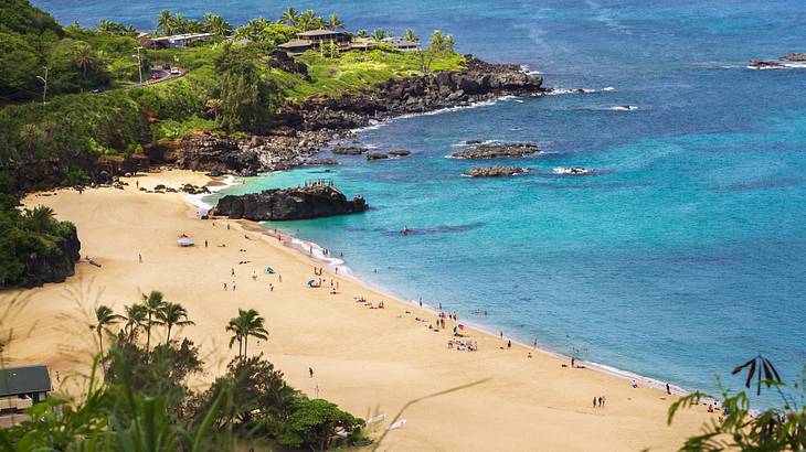 A sandy beach with blue water and greenery and rocks surrounding the sand