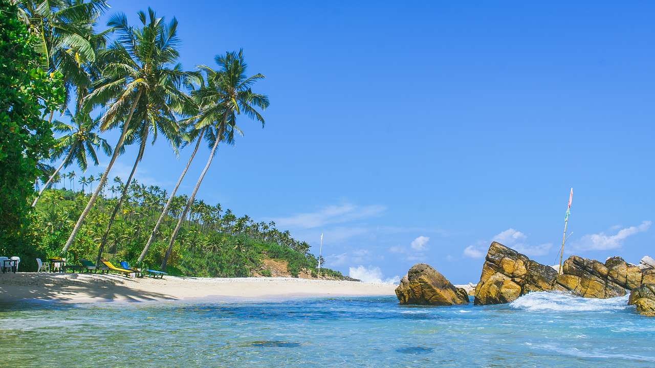 An empty beach with chairs, palm trees, clear waters, and some rocks near the shore