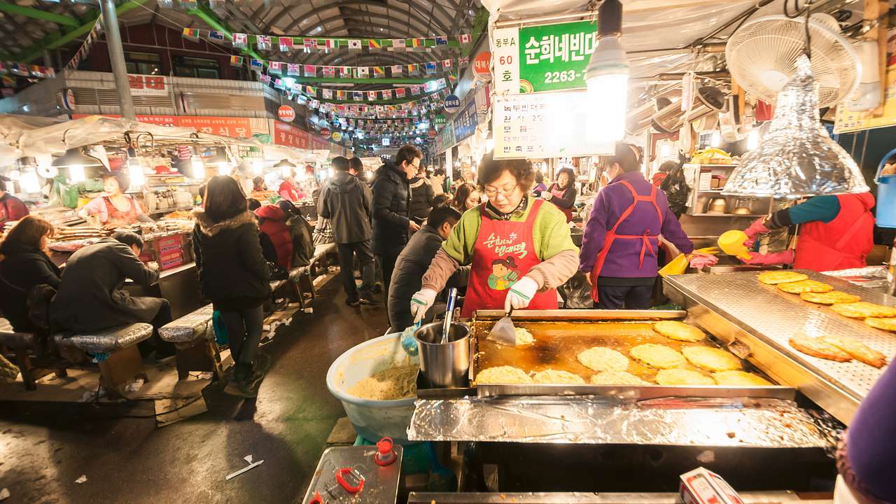 A woman cooking pancakes on a grill amidst people and other stalls selling food