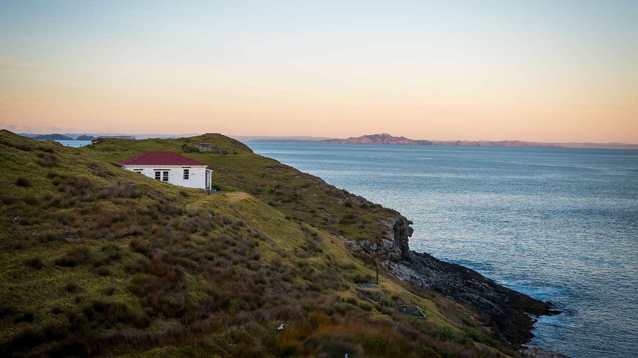 A white house nestled amidst a green mountain beside the blue ocean at dusk