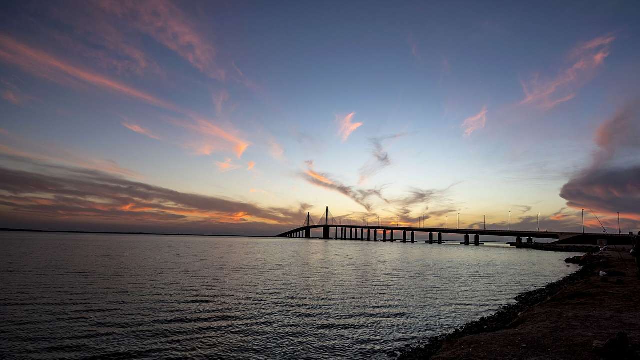 A beach with a distant view of a long bridge at sunset