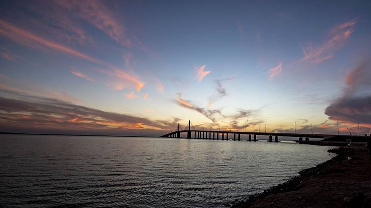 A beach with a distant view of a long bridge at sunset