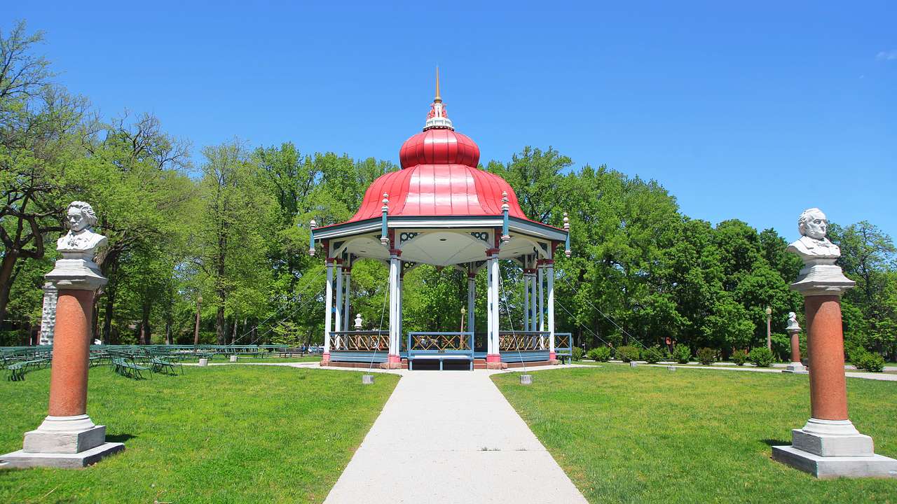 A walkway through the grass to a gazebo with a red roof and green trees behind it
