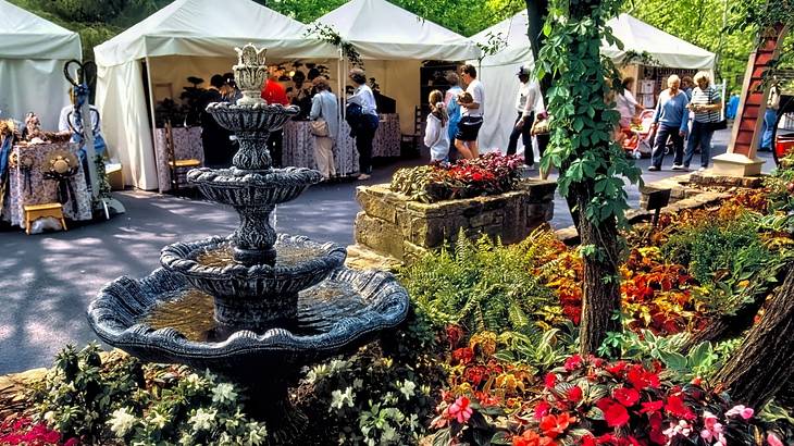 A small fountain between colorful flowers against crowds wandering tented stalls