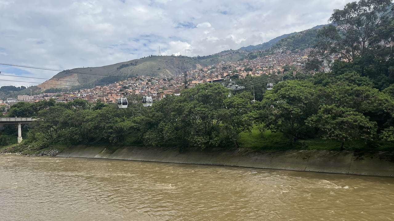 Looking across a brown river to trees and homes up a mountainside with a cable car