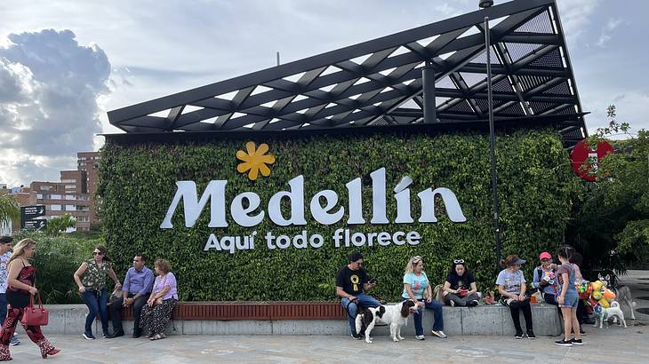 People sitting along a green wall that says "Medellin Aqui todo florece"