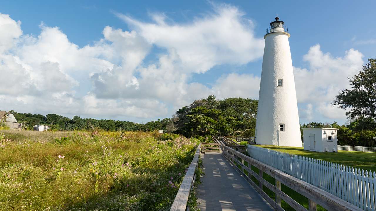 A wooden path leading to a white lighthouse and a shed underneath a partly cloudy sky