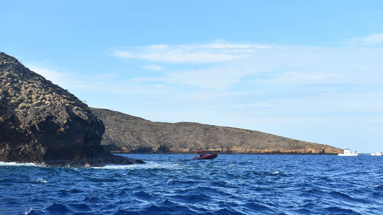 A red boat on the ocean with rocks to the side under blue sky