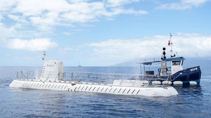 A white submarine on the surface of the ocean beside a blue and white tugboat