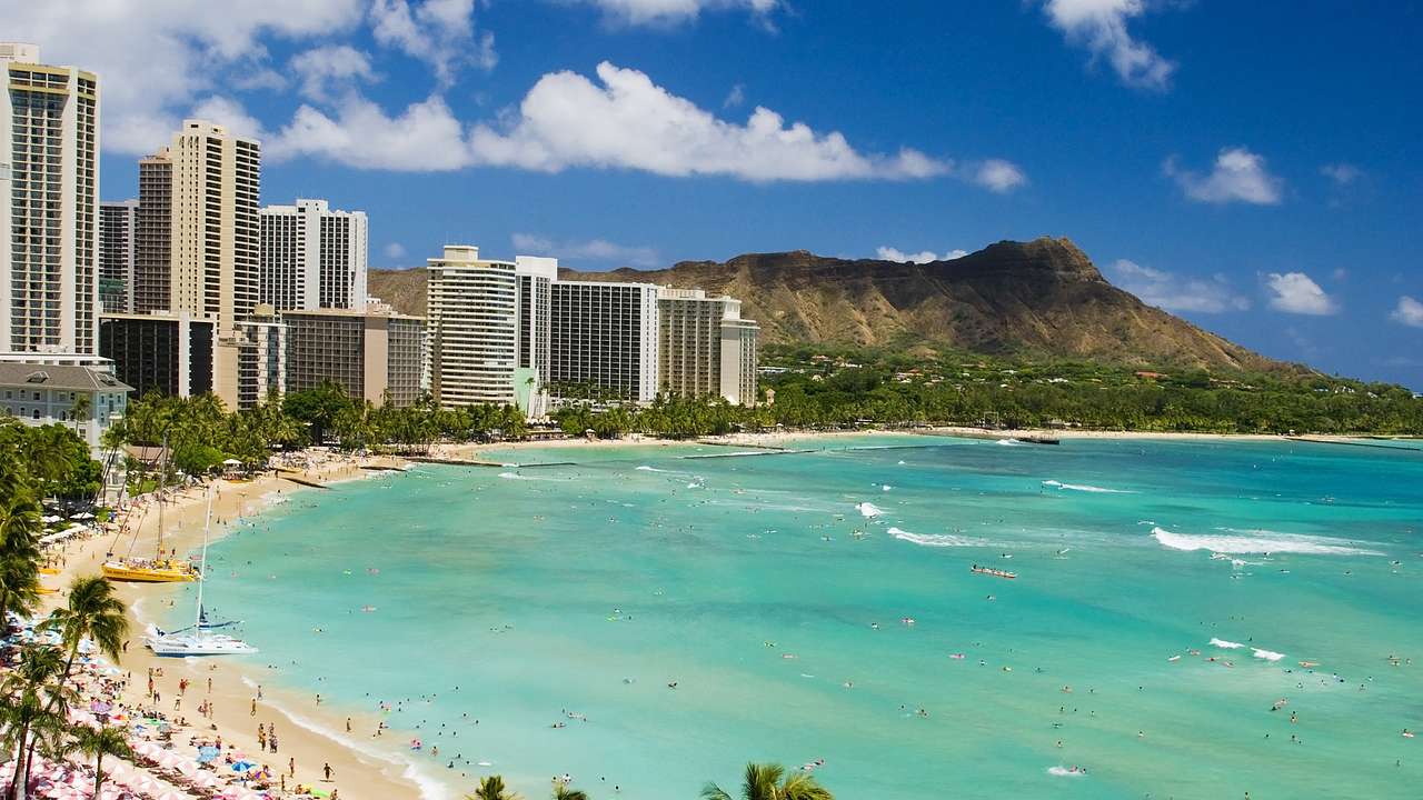 Crystal blue ocean with a beach, palm trees, buildings, and a mountain to the side