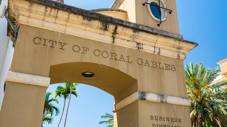 Archway with text above, against a blue sky, surrounded by palm trees