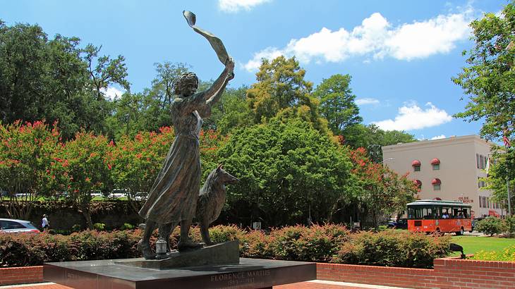 A statue of a woman and a dog, surrounded by trees, bushes, and a building