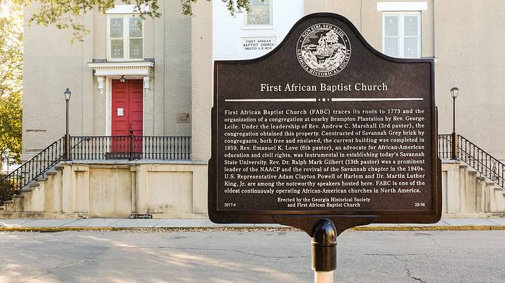 A placard with writings on it, with a cream-colored building with a red door behind