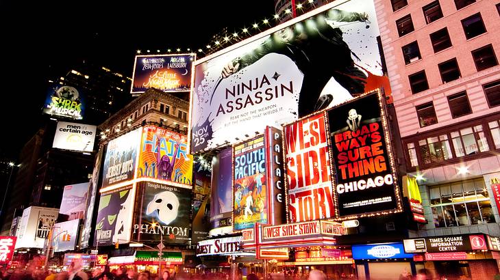 A building with lit-up billboards for broadway shows with people walking in front