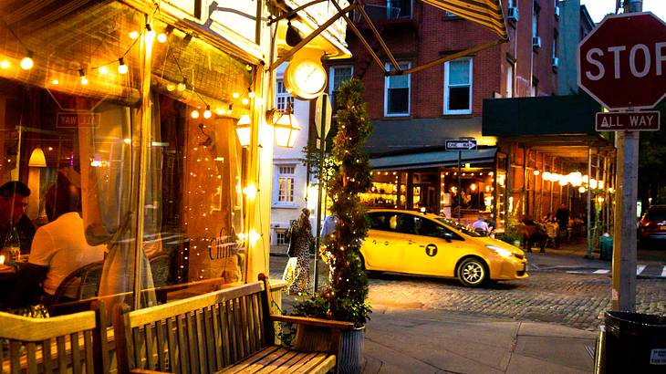 The front of a building with fairy lights next to a bench and a street with a taxi