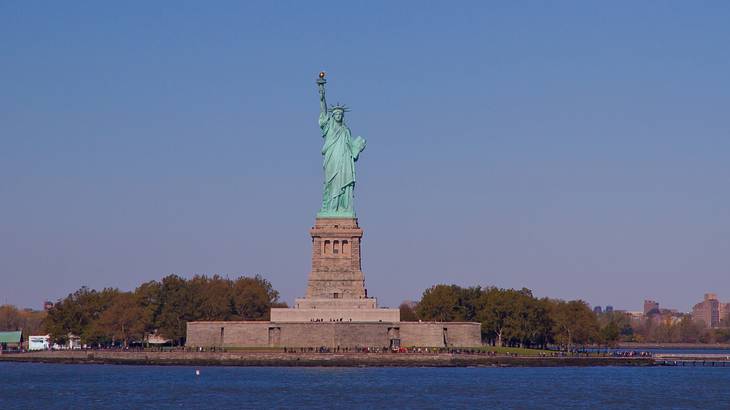 The Statue of Liberty with water surrounding and trees behind it under evening sky