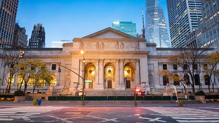 A stone library building at night with a road in front and skyscrapers behind it