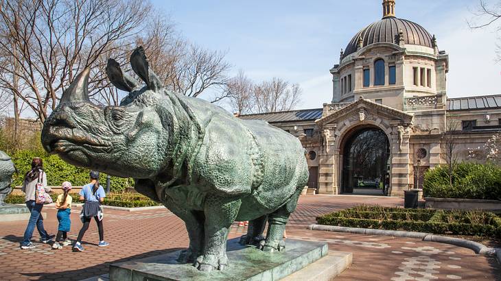 A statue of a rhino in front of an old building with three people walking towards it