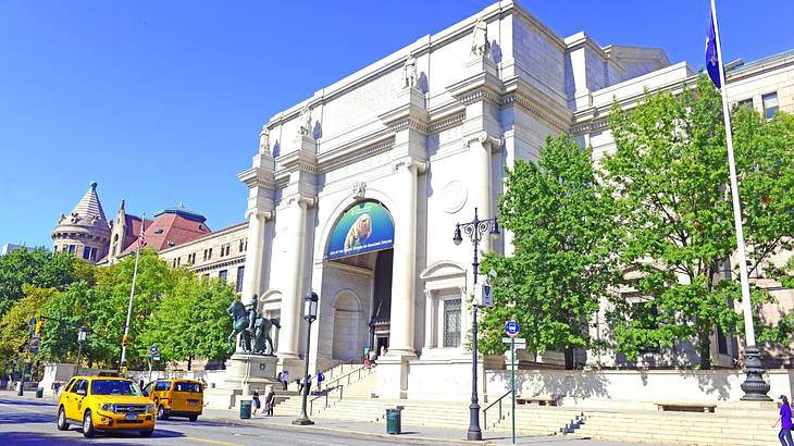 A white stone museum building with a road, yellow taxis, and trees in front of it