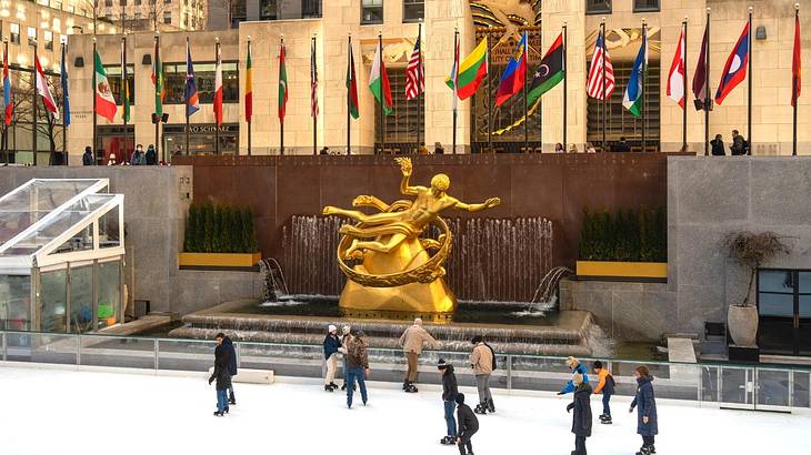 An ice rink with people skating with a statue, flags, and buildings behind it