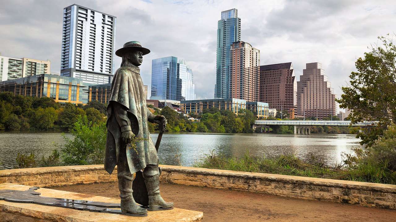 A statue of a man holding a guitar near a river with a city skyline in the background