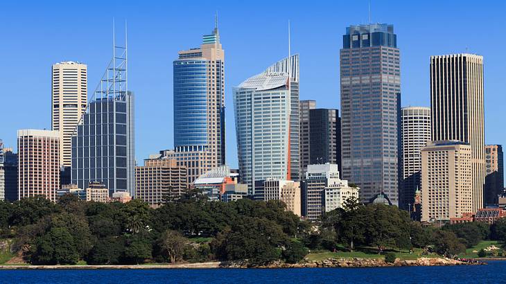 A city skyline of tall skyscrapers near trees and a body of water