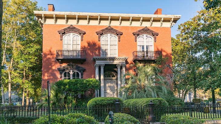 A red brick house with a large window, surrounded by a walkway, trees, and bushes