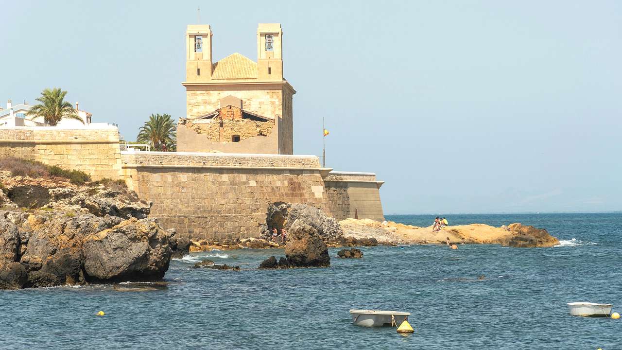 A small stone castle surrounded by water with rocks and two small anchored boats