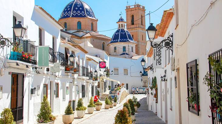 A Spanish town street with white buildings, plants, and a church with blue domes