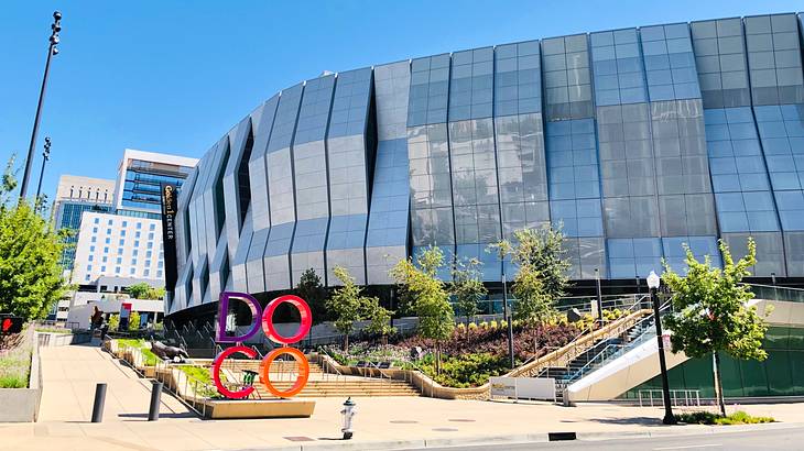 A large, curved building with angled exterior panels and greenery in front of it