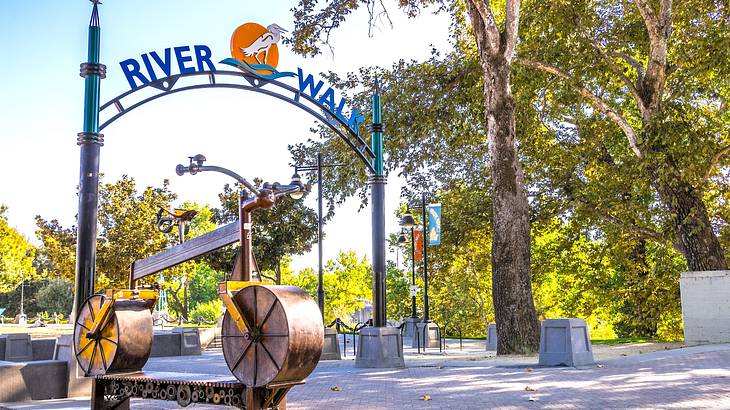 A metal arch with a "River Walk" sign over a path next to a metal sculpture and trees