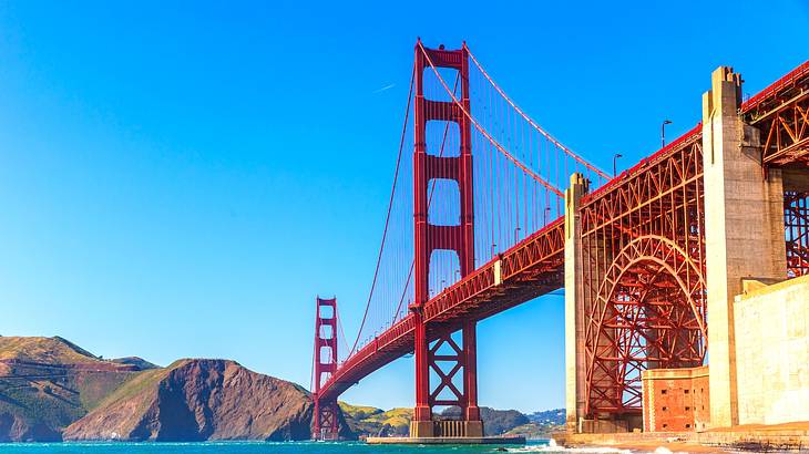 A large red suspension bridge over the water with small hills on one side